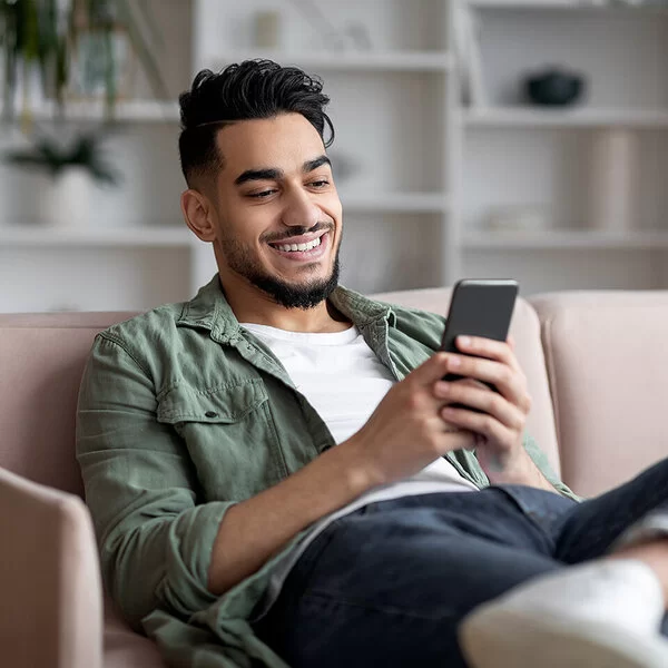 Ein junger Mann sitzt gemütlich zuhause auf dem Sofa und spielt auf dem Smartphone. Er lacht, es macht ihm Spaß.