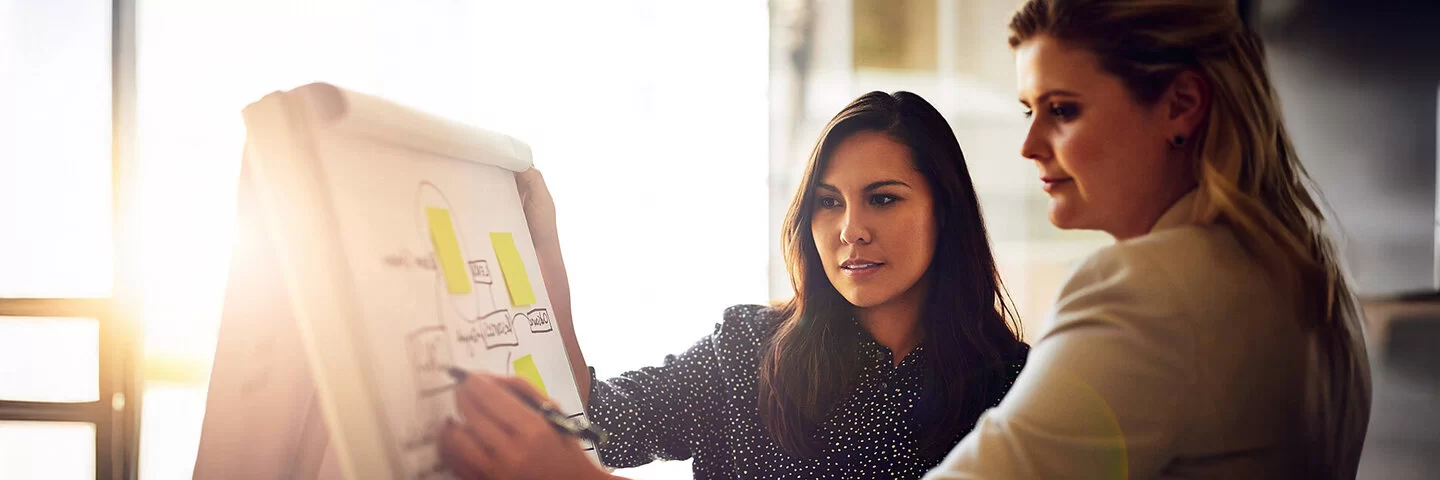 Zwei junge Frauen stehen vor einem Flipchart. Die eine schreibt mit einem Stift etwas in ein Kästchen eines Diagramms. Die andere liest das Geschriebene.