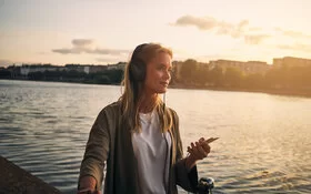 Eine junge Frau steht an einem Flussufer und hört mit Kopfhörern einen Podcast.