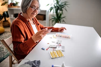 Eine ältere Frau mit weißgrauen Haaren, mit Brille und einer rotbraunen Bluse sitzt an einem weißen Tisch und hält einen Blister mit Tabletten in der rechten Hand. Vor ihr liegen weitere Medikamente auf dem Tisch verteilt. Im Hintergrund ist ein Wohnzimmer zu sehen.