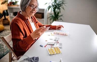 Eine ältere Frau mit weißgrauen Haaren, mit Brille und einer rotbraunen Bluse sitzt an einem weißen Tisch und hält einen Blister mit Tabletten in der rechten Hand. Vor ihr liegen weitere Medikamente auf dem Tisch verteilt. Im Hintergrund ist ein Wohnzimmer zu sehen.