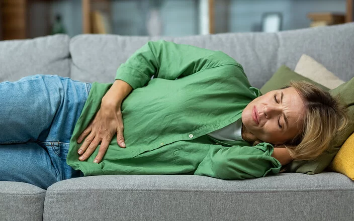 Eine Frau in den Dreißigern liegt mit Schmerz ausdrückendem Gesicht und geschlossenen Augen auf einem Sofa und greift sich mit der rechten Hand an den Unterbauch.