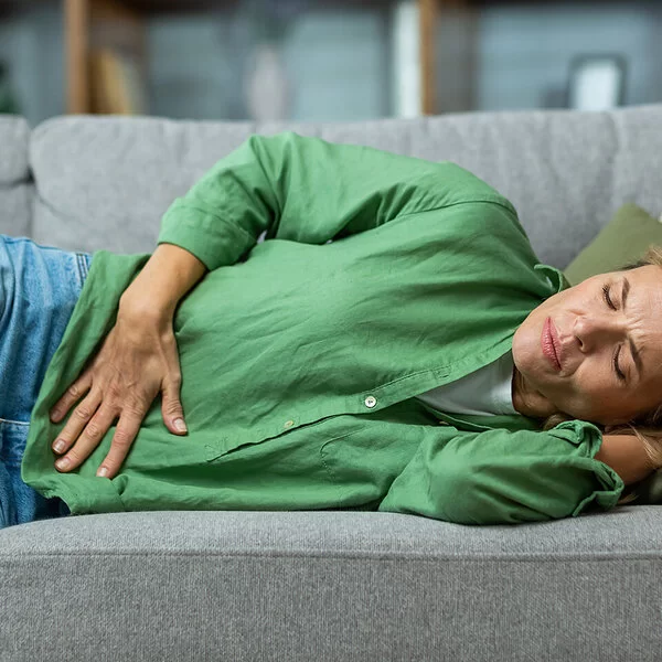 Eine Frau in den Dreißigern liegt mit Schmerz ausdrückendem Gesicht und geschlossenen Augen auf einem Sofa und greift sich mit der rechten Hand an den Unterbauch.