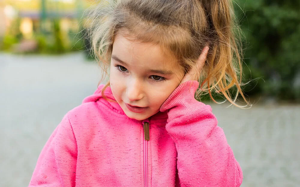 Ein ungefähr vier Jahre altes Mädchen mit rosa Kapuzenpullover und blondem Zopf hält sich eine flache Hand gegen ihr linkes Ohr.