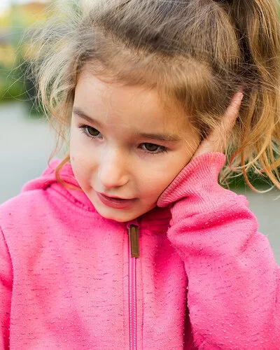 Ein ungefähr vier Jahre altes Mädchen mit rosa Kapuzenpullover und blondem Zopf hält sich eine flache Hand gegen ihr linkes Ohr.