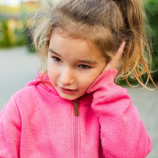 Ein ungefähr vier Jahre altes Mädchen mit rosa Kapuzenpullover und blondem Zopf hält sich eine flache Hand gegen ihr linkes Ohr.