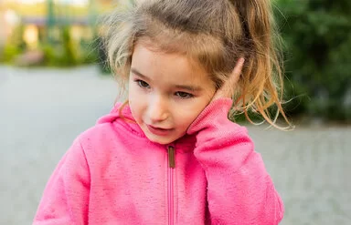 Ein ungefähr vier Jahre altes Mädchen mit rosa Kapuzenpullover und blondem Zopf hält sich eine flache Hand gegen ihr linkes Ohr.