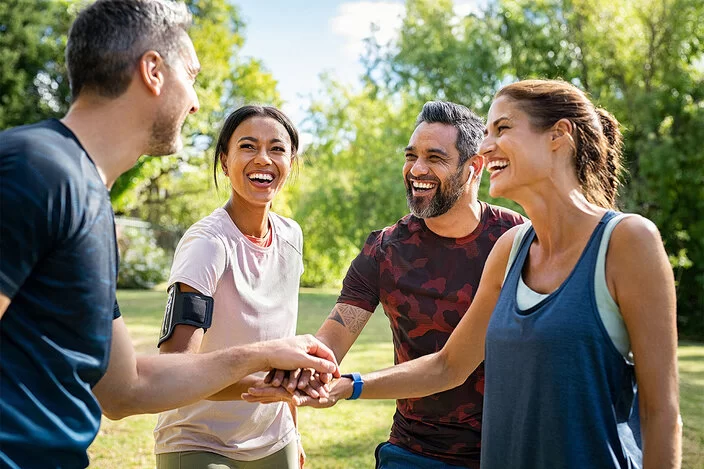 Eine Gruppe junger Erwachsener in Sportkleidung steht lächelnd im Kreis im Park und legt die Hände übereinander in die Mitte.