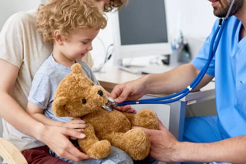 Ein kleiner Junge mit einem Teddybär sitzt auf dem Schoß seiner Mutter, während ein Arzt mit einem Stethoskop so tut, als würde er die Lungen des Teddybären abhören.