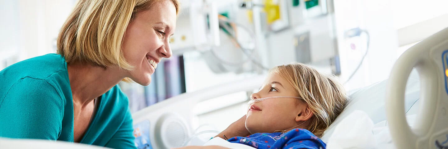 Eine junge Frau beugt sich lächelnd zu einem Kind, das in einem Krankenhausbett liegt.