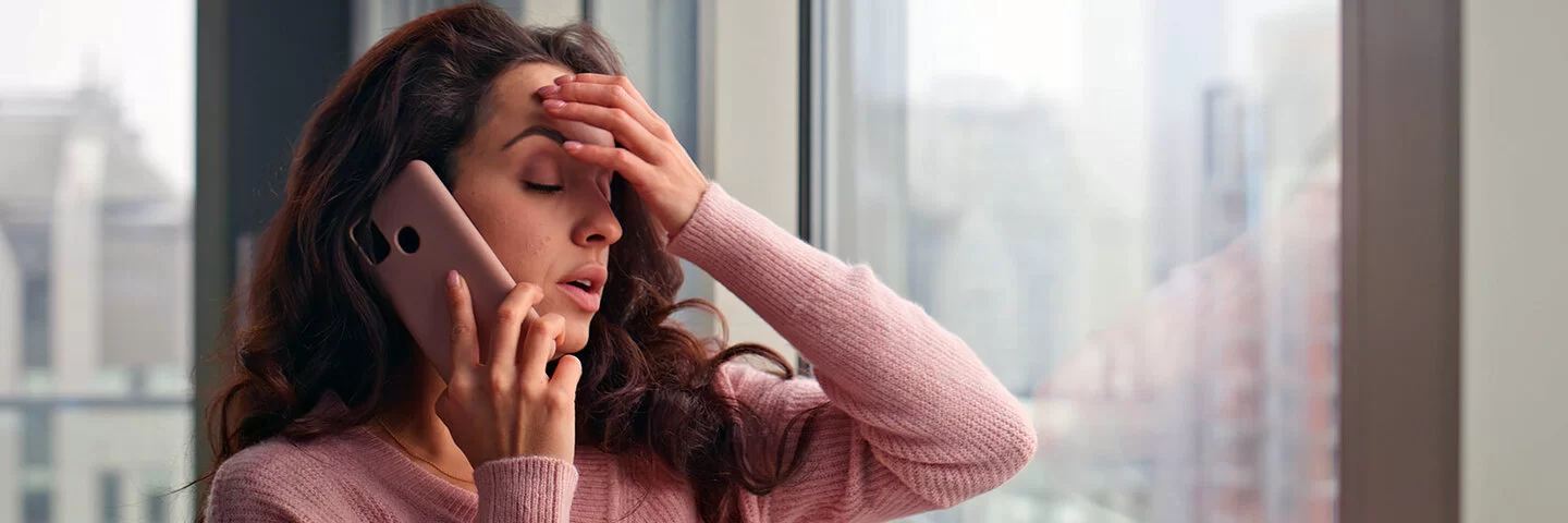 Eine gestresst wirkende junge Frau telefoniert mit ihrem Smartphone am Fenster eines Büros oder einer Wohnung. Ihre Augen sind geschlossen. Die rechte Hand hält das Telefon, mit der linken Hand greift sie sich an die Stirn.