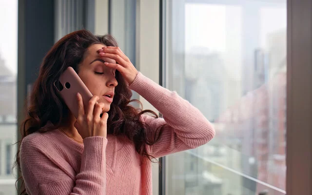 Eine gestresst wirkende junge Frau telefoniert mit ihrem Smartphone am Fenster eines Büros oder einer Wohnung. Ihre Augen sind geschlossen. Die rechte Hand hält das Telefon, mit der linken Hand greift sie sich an die Stirn.