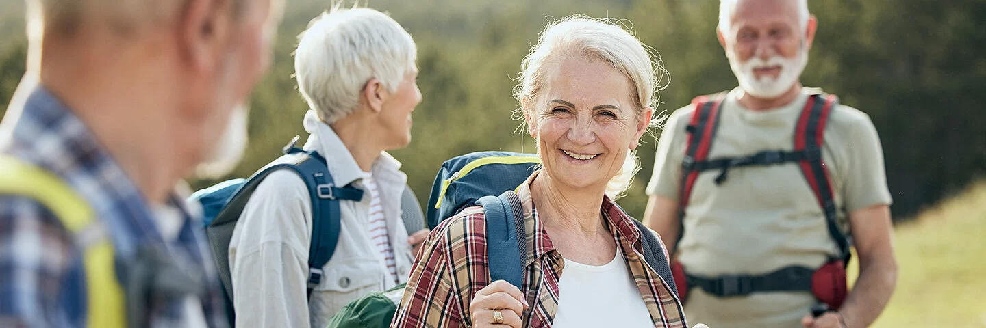 Zwei ältere Frauen und zwei ebenfalls ältere Männer sind wandern. Sie stehen an einem grünen Hang, tragen leichte Wanderrucksäcke auf den Schultern und lächeln.