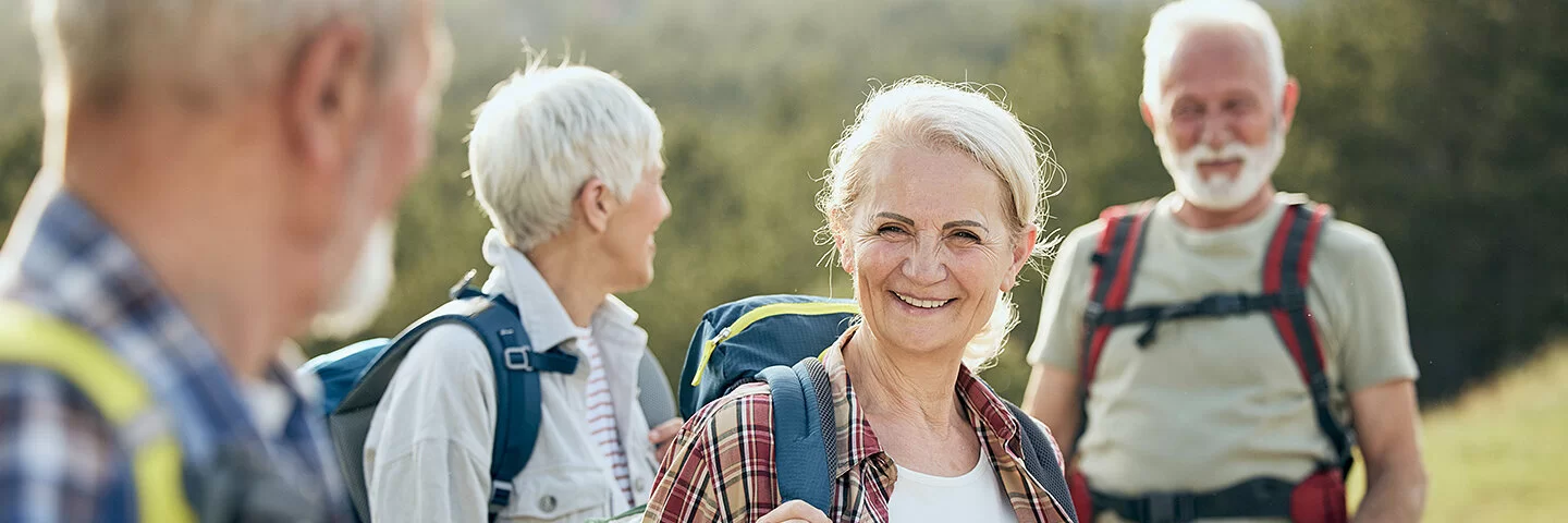 Zwei ältere Frauen und zwei ebenfalls ältere Männer sind wandern. Sie stehen an einem grünen Hang, tragen leichte Wanderrucksäcke auf den Schultern und lächeln.