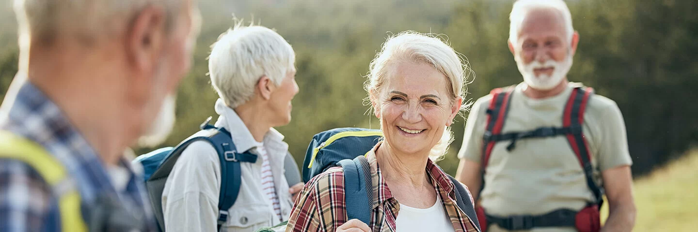 Zwei ältere Frauen und zwei ebenfalls ältere Männer sind wandern. Sie stehen an einem grünen Hang, tragen leichte Wanderrucksäcke auf den Schultern und lächeln.