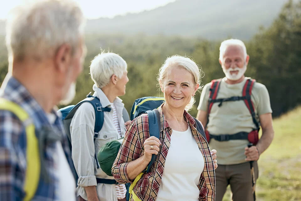 Zwei ältere Frauen und zwei ebenfalls ältere Männer sind wandern. Sie stehen an einem grünen Hang, tragen leichte Wanderrucksäcke auf den Schultern und lächeln.