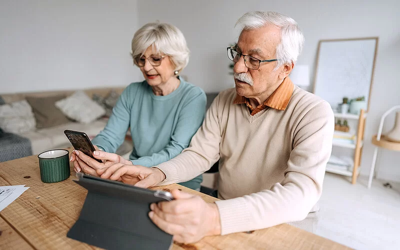 Ein Seniorenpaar sitzt im Wohnzimmer am Esstisch. Die Frau hält ein Smartphone in der Hand und der Mann bedient ein auf dem Tisch stehendes Tablet.