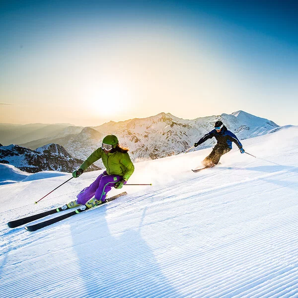 Ein Mann und eine Frau fahren auf Skiern rasant eine Piste hinab. Im Hintergrund sind schneebedeckte Berge zu sehen.