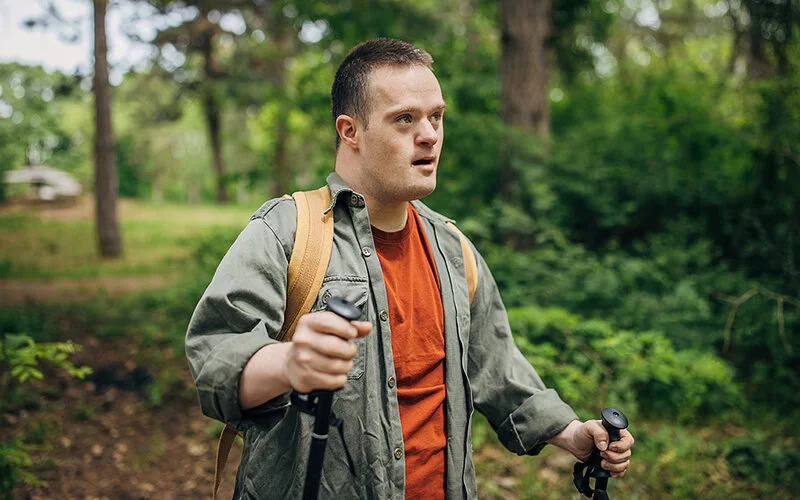 Ein junger Mann mit Down-Syndrom wandert mit Nordic-Walking-Stöcken und Rucksack durch einen Wald.