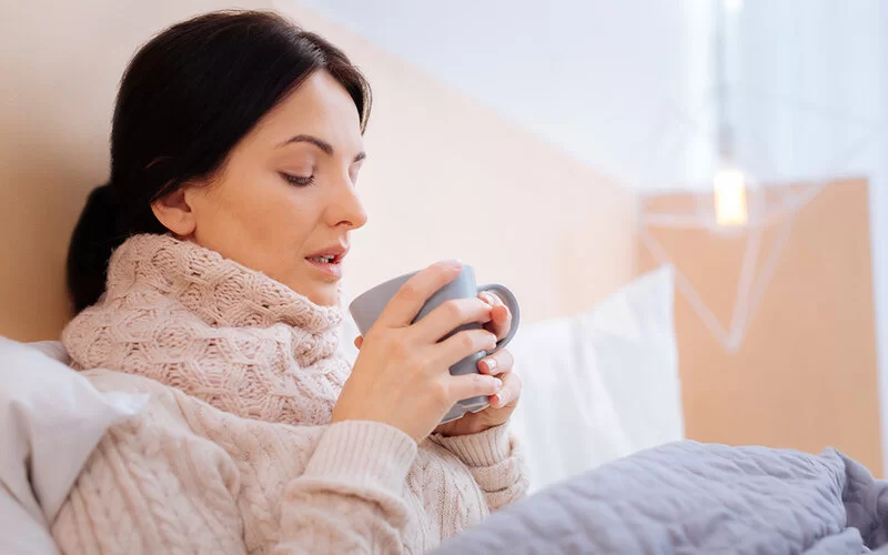 Junge Frau mit warmem Pullover, zugedeckt im Bett. In der Hand hält sie eine Tasse mit heißem Tee.