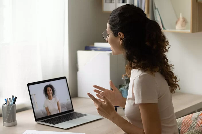 Eine junge Frau mit dunklen, lockigen Haaren und Brille, im beigen T-Shirt, sitzt am Schreibtisch und unterhält sich per Video-Chat mit einer anderen Frau.