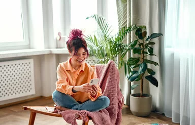 Eine junge Frau mit zusammengebundenen Haaren sitzt in einem orangefarbenen Hemd und Jeans im Schneidersitz auf einem großen Holzstuhl mit einer pinken Decke im Wohnzimmer. Sie schaut lächelnd auf ihr Smartphone.