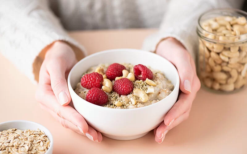 Die Hände einer Frau halten auf einem Tisch eine Porzellanschüssel mit Haferbrei, der mit Cashewkernen und Himbeeren garniert ist.