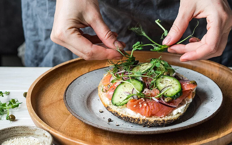 Nahaufnahme eines Tellers, auf dem ein Brötchen mit Lachs liegt. Darauf befinden sich Gurkenscheiben.