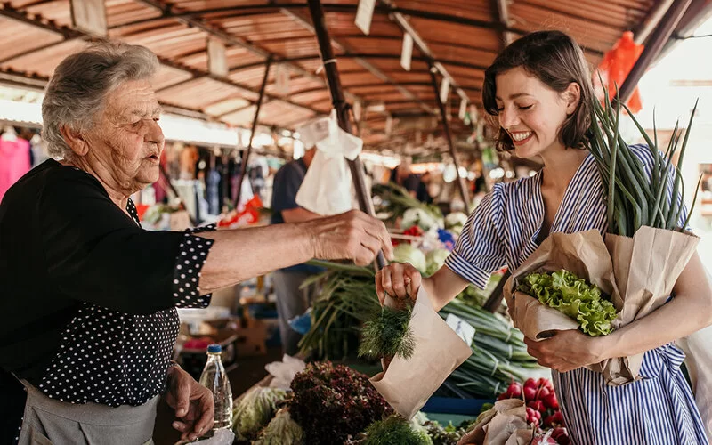 Frau wird beim Einkauf von alter Händlerin am Wochenmarkt beraten und lächelt.