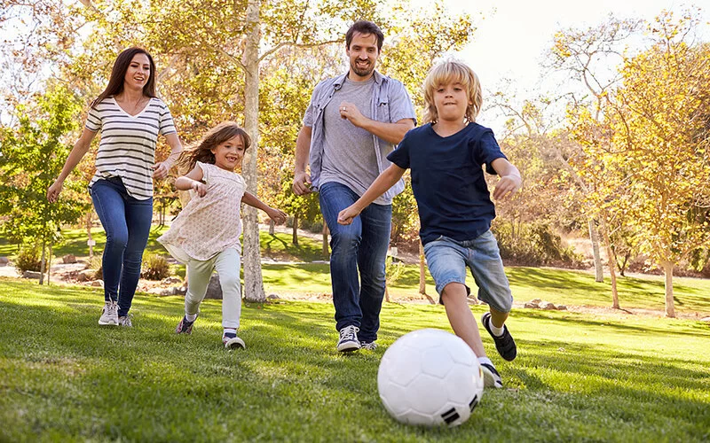 Eine Mutter und ein Vater spielen bei schönem Wetter mit ihrer Tochter und ihrem Sohn Fußball in einem Park. Sie lachen und haben viel Spaß dabei.