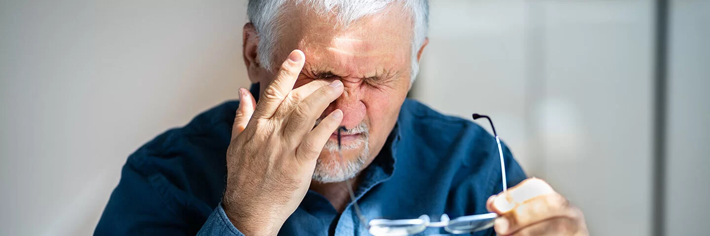 Ein älterer Mann hält in der einen Hand die Brille und fasst sich mit dem Finger seiner anderen Hand ins Auge.