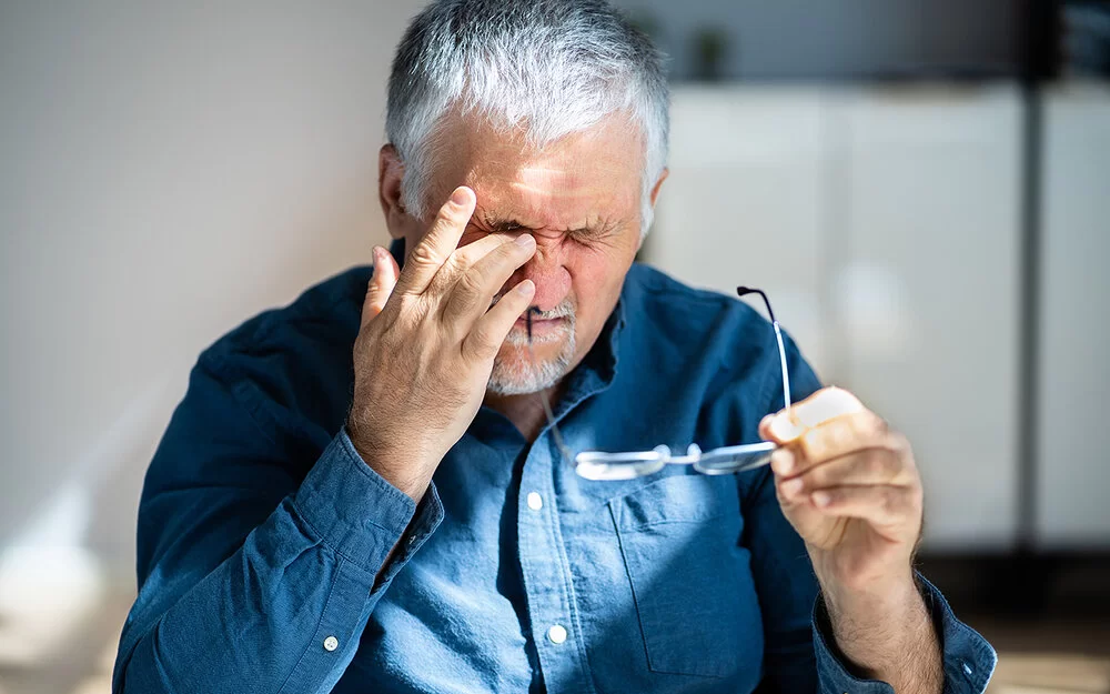 Ein älterer Mann hält in der einen Hand die Brille und fasst sich mit dem Finger seiner anderen Hand ins Auge.