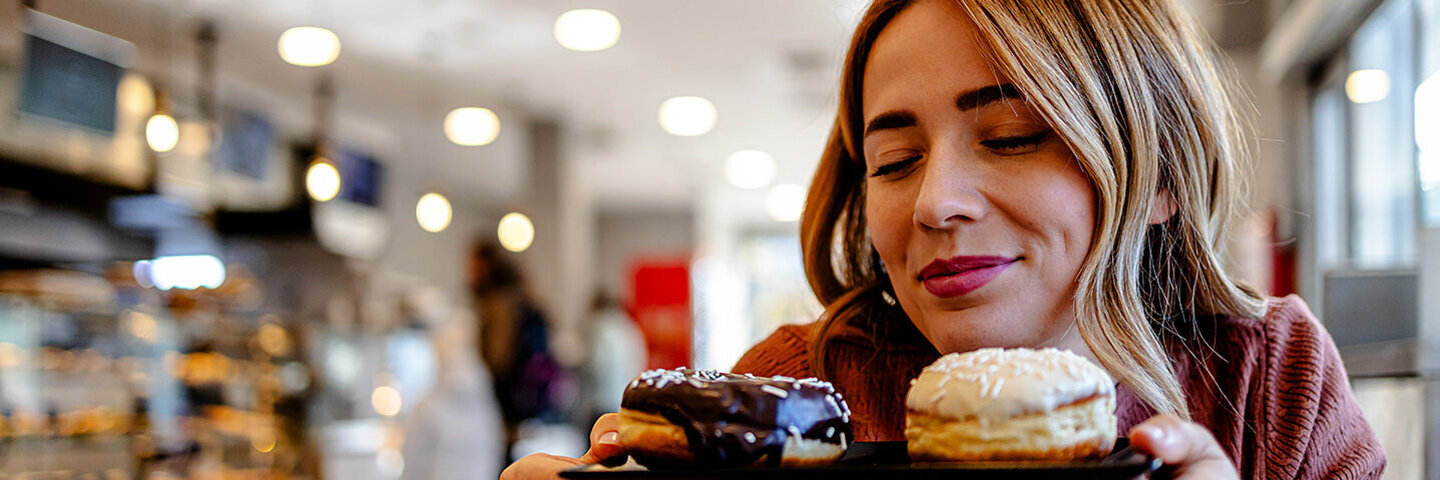 Eine Frau mit langen blondgesträhnten Haaren sitzt in einem Café, hält ein kleines Tablett mit Donuts in den Händen und hat dabei genussvoll die Augen geschlossen.