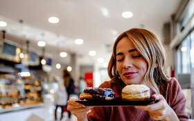 Eine Frau mit langen blondgesträhnten Haaren sitzt in einem Café, hält ein kleines Tablett mit Donuts in den Händen und hat dabei genussvoll die Augen geschlossen.