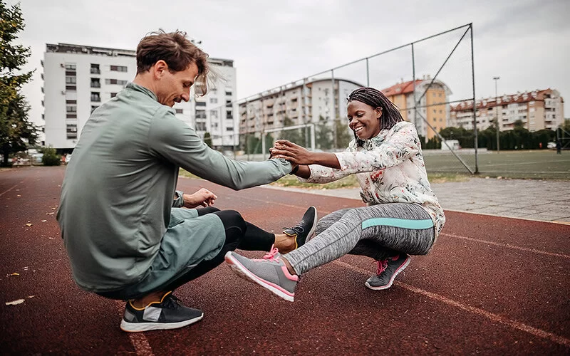 Auf der Tartanbahn einer Sportanlage in einem Wohngebiet mit mehrstöckigen Häusern macht ein junges Paar Dehnübungen. Der Mann und die Frau knien dabei, fassen sich an den Händen und strecken jeweils ihr linkes Bein aus.