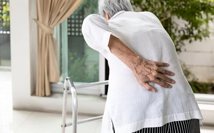 Rückenansicht einer älteren Frau mit Rundrücken. Vor der Frau, die sich mit der rechten Hand in die Lendengegend greift, steht eine Gehhilfe.