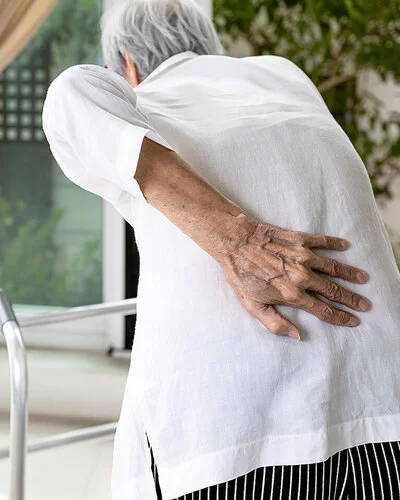 Rückenansicht einer älteren Frau mit Rundrücken. Vor der Frau, die sich mit der rechten Hand in die Lendengegend greift, steht eine Gehhilfe.