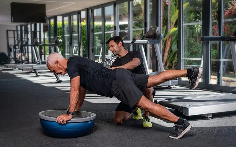 Ein Mann stützt sich unter Anleitung eines Trainers auf ein Sportgerät und hebt dabei ein Bein.