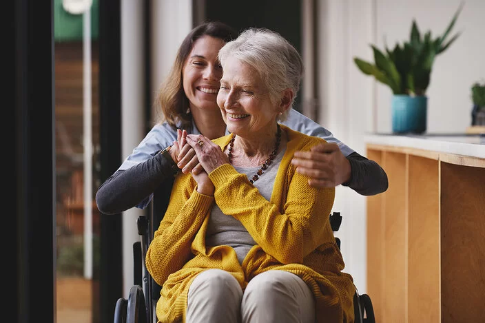 Eine Seniorin mit kurzen weißen Haaren und einer gelben Strickjacke sitzt im Rollstuhl und hält lächelnd die Hand der Pflegerin hinter sich.