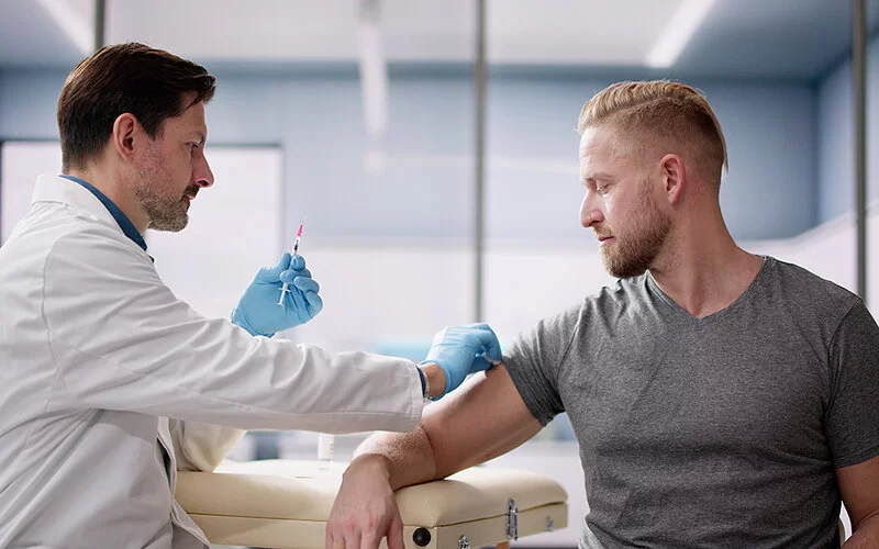 Junger Mann in grauem T-Shirt in einem Untersuchungszimmer. Der Arzt im weißen Kittel hält eine Spritze in der Hand und desinfiziert dem Patienten die Haut am Oberarm.