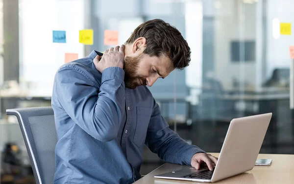 Ein dunkelhaariger Mann in einem blauen Hemd sitzt mit gebeugtem Kopf an einem Schreibtisch vor einem Laptop. Er fasst sich mit einer Hand an den schmerzenden Nacken. Im Hintergrund ist die Glaswand eines Büros zu sehen.