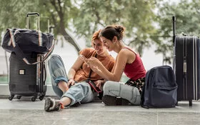 Zwei junge Frauen in sommerlicher Kleidung sitzen auf dem Boden eines Gebäudes. Sie schauen lachend auf ein Handy, das eine von ihnen in der Hand hält. Um sie herum stehen zwei Reisekoffer, eine Reisetasche und ein Rucksack.