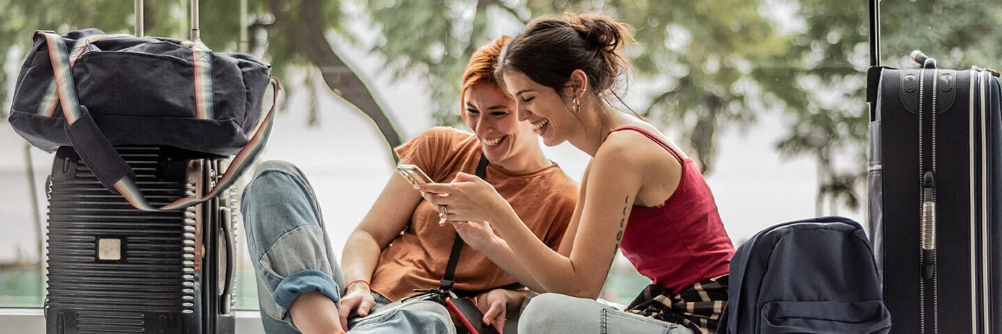 Zwei junge Frauen in sommerlicher Kleidung sitzen auf dem Boden eines Gebäudes. Sie schauen lachend auf ein Handy, das eine von ihnen in der Hand hält. Um sie herum stehen zwei Reisekoffer, eine Reisetasche und ein Rucksack.