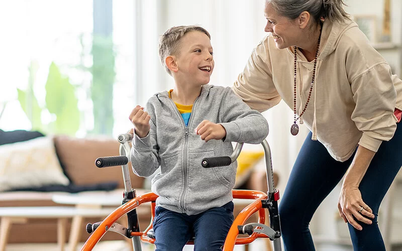 Ein Junge mit eingeschränkter Beinbeweglichkeit sitzt in einem speziellen physiotherapeutischen Stuhl. Neben ihm steht seine Physiotherapeutin, die mit ihrem rechten Arm an die Stuhllehne greift. Beide schauen sich an und lächeln.