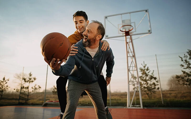 Ein Vater spielt mit seinem Sohn Basketball auf einem Outdoor-Basketballplatz. Er trägt seinen Sohn huckepack und hält den Ball mit ausgestrecktem rechten Arm in der Hand.