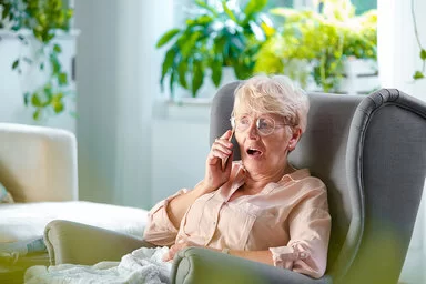 Ältere Frau sitzt mit einer Decke auf den Beinen in einem Sessel und telefoniert.