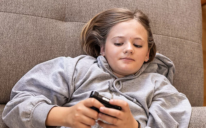 Ein Mädchen im Teenageralter liegt auf einem grauen Sofa. Es ist nur ihr Oberkörper zu sehen. Sie hält ein Blutzuckermessgerät in der Hand und macht einen zufriedenen Eindruck.