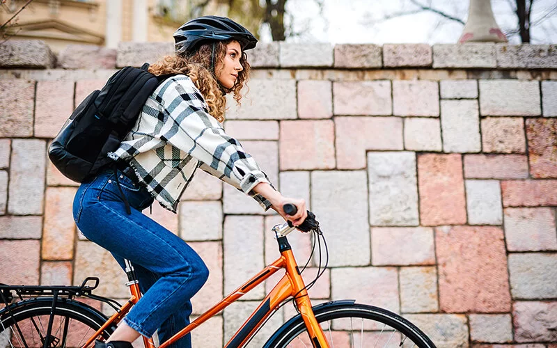 Eine junge Frau in Freizeitkleidung auf einem Fahrrad. Sie trägt einen Helm und einen schwarzen Rucksack und fährt an einer Steinmauer entlang.