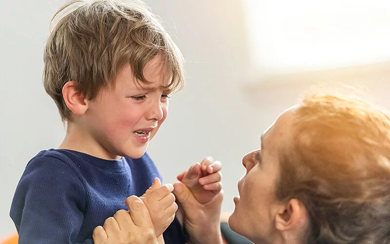 Aufnahme eines weinenden Kindes, das einen blauen Pullover trägt. Vorn im Bild ist der Kopf einer erwachsenen Person zu sehen, die zu ihm aufblickt. Die Person umfasst die Handgelenke des Kindes und scheint auf es einzureden.