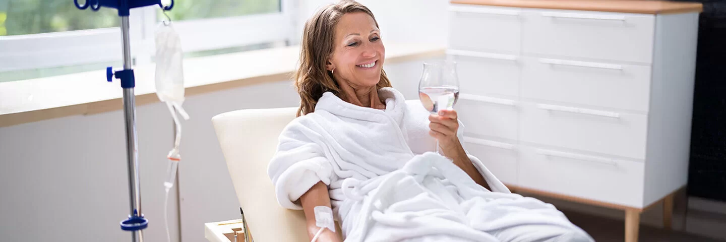 Eine Frau liegt im weißen Bademantel auf einer Liege. Über einen Tropf erhält sie eine Infusion in ihren rechten Arm. In der linken hält sie ein großes Glas Wasser, das sei freudestrahlend anschaut.